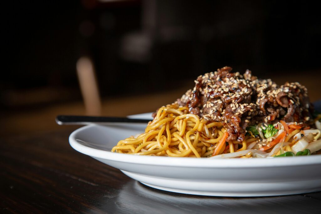 A close-up of Asian noodles with beef and vegetables, garnished with sesame seeds on a white plate.